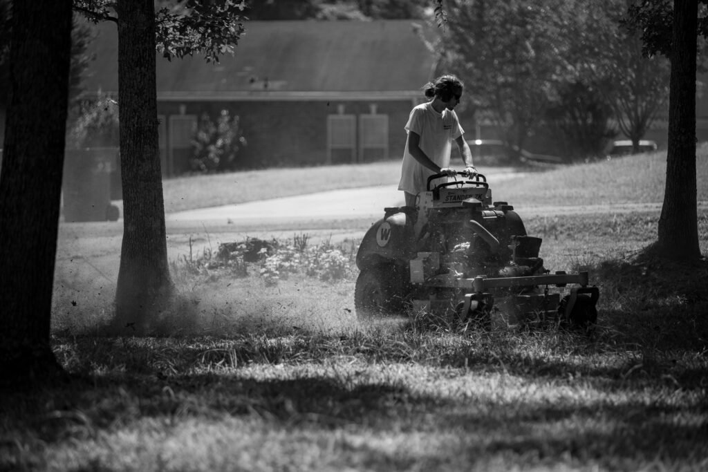 A person operating a lawnmower in a shaded, grassy yard, in black and white.